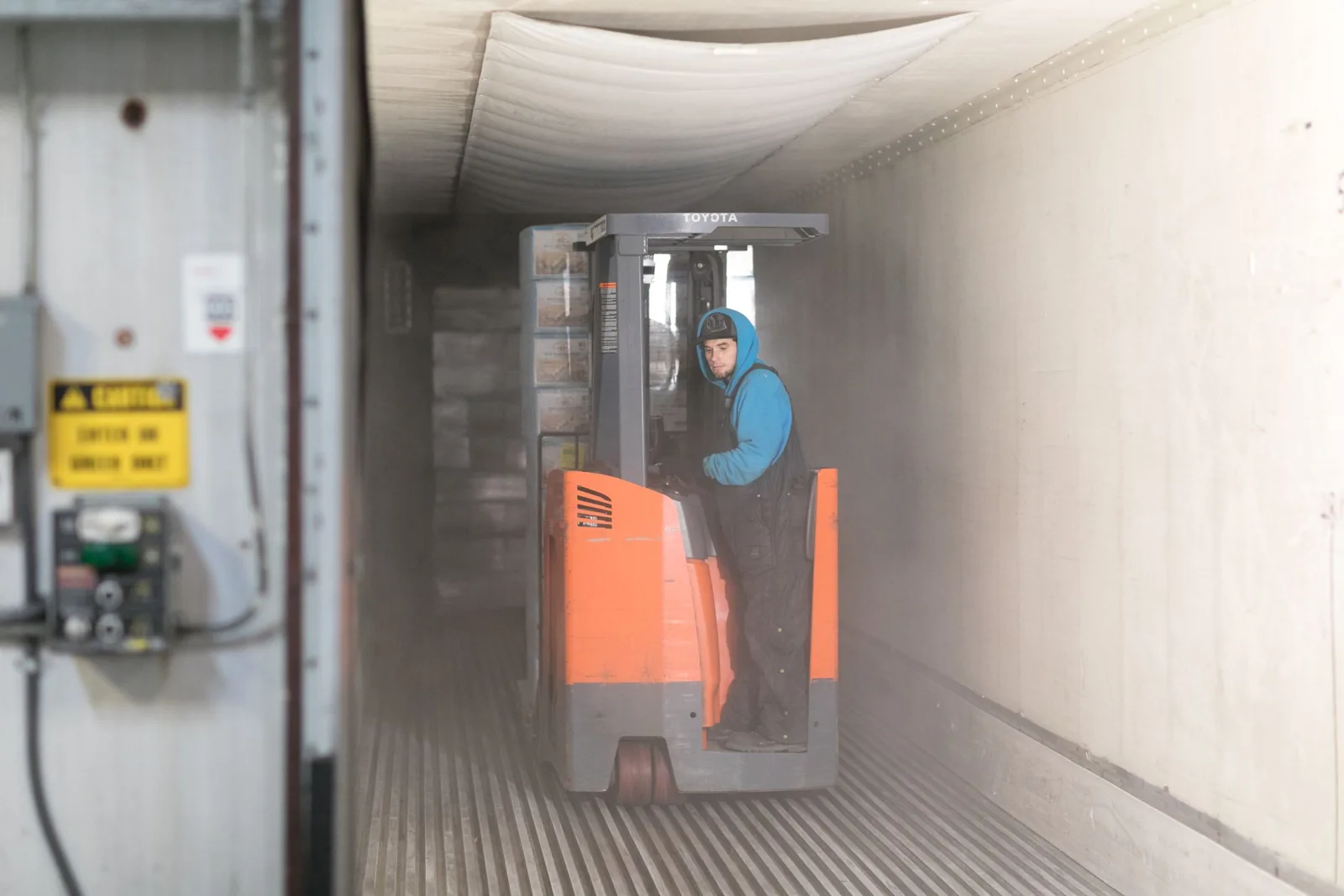CWI warehouse worker in cold-weather gear operating a reach forklift inside a trailer loaded with pallets